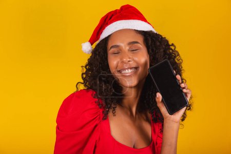 Téléchargez les photos : Souriant jeune Santa femme en chapeau de Noël tenant téléphone portable avec écran vide vide isolé sur fond jaune. - en image libre de droit