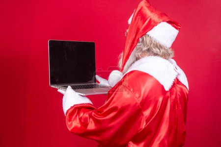 Photo for Santa Claus using a laptop on red background with his back turned. - Royalty Free Image