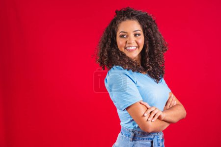 Photo for Beauty portrait of a Brazilian woman with afro hairstyle and glamour makeup. Latin woman. Mixed race. Curly hair. Hair style. Red background. Cross arms - Royalty Free Image