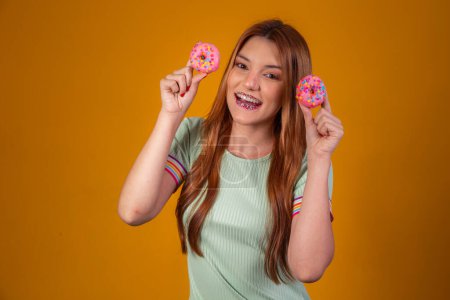 Foto de Chica con estilo con el pelo largo positivamente posa, sosteniendo donas rosadas frescas con polvo listo para disfrutar de dulces. - Imagen libre de derechos