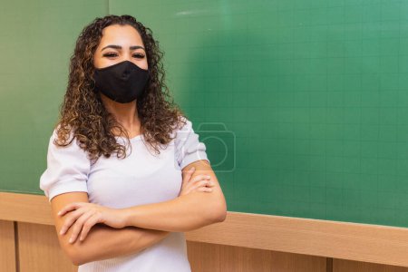 Téléchargez les photos : Jeune enseignante avec les bras croisés en classe avec tableau noir en arrière-plan portant un masque chirurgical dans la nouvelle normale. Concept de retour à l'école après une pandémie - en image libre de droit