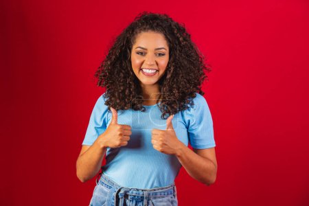 Photo for Beauty portrait of a Brazilian woman with afro hairstyle and glamour makeup. Latin woman. Mixed race. Curly hair. Hair style. Red background. Thumbs up. - Royalty Free Image