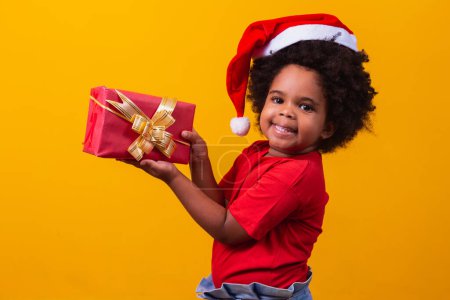 Téléchargez les photos : Enfant afro souriant au chapeau rouge du Père Noël tenant à la main un cadeau de Noël. Concept de Noël. - en image libre de droit