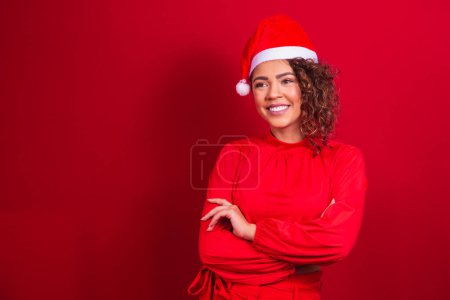 Foto de Mujer afro con sombrero de Navidad de pie sobre fondo rojo con los brazos cruzados. retrato aislado. - Imagen libre de derechos