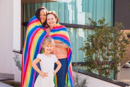 Téléchargez les photos : Couple lesbien souriant avec leur fille adoptive. gay famille avec lgbt drapeau - en image libre de droit
