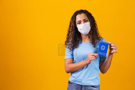 Foto de Mujer afro con máscara quirúrgica que sostiene la tarjeta de trabajo brasileña sobre fondo amarillo. Trabajo, economía y concepto pandémico - Imagen libre de derechos