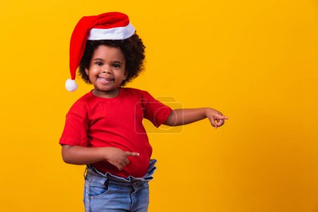 Téléchargez les photos : Petite Afro fille en T-shirt rouge et chapeau de Père Noël habillé pour Noël avec espace pour le texte pointant. - en image libre de droit