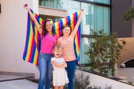 Téléchargez les photos : Couple lesbien souriant avec leur fille adoptive. gay famille avec lgbt drapeau - en image libre de droit