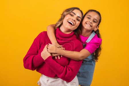 Photo for Photo of a beautiful young mommy holding her arms hugging her little daughter good mood adorable sincere heart feelings in the back jeans t-shirts isolated with yellow color background - Royalty Free Image