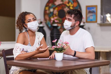 Foto de Un par de amigos de seguridad hablando sentados en un restaurante al aire libre con máscara de seguridad contra la pandemia de coronavirus covid-19. Nuevo concepto de vida normal - Imagen libre de derechos