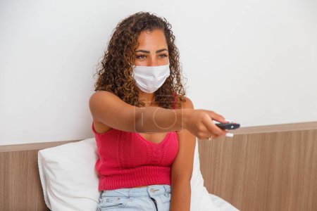Photo for Woman at home isolated in pandemic wearing surgical mask and watching TV. - Royalty Free Image