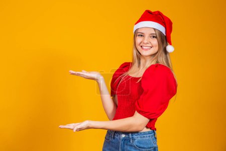 Photo for Positive girl in Santa hat pointing at text space. Promotion and advertisements isolated on yellow background. - Royalty Free Image