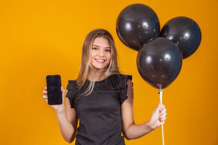 Téléchargez les photos : Charmante jeune femme souriante tenant un téléphone portable avec écran vide sur fond jaune avec des ballons à air portrait studio. Règlement du vendredi noir - en image libre de droit