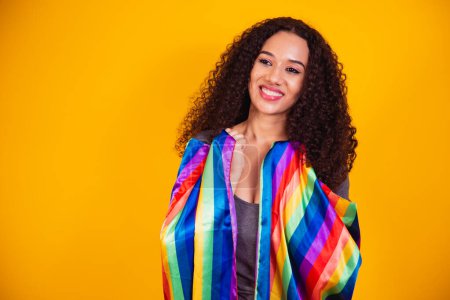 Photo for Young curly hair woman covering with lgbt pride flag. Alone. One. Keeping fist up, covering LGBT flag. LGBT+ flag on yellow background. - Royalty Free Image