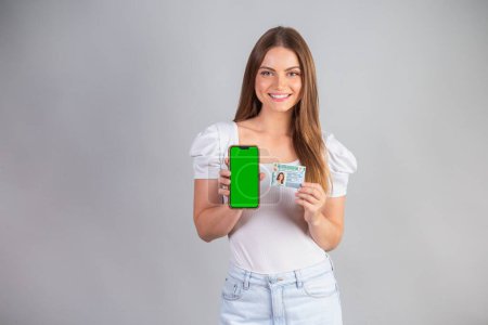 Photo for Blonde Brazilian woman holding a motor vehicle driver's license and smartphone. Translation in English (national driver's license) - Royalty Free Image