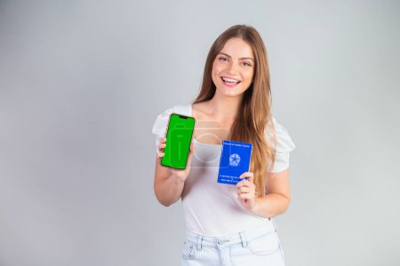 Téléchargez les photos : Femme brésilienne blonde tenant une carte nationale de travail et de sécurité sociale et un smartphone. Traduction en anglais (carte d'emploi et sécurité sociale) - en image libre de droit