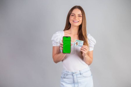 Photo for Blonde Brazilian woman holding a motor vehicle driver's license and smartphone. Translation in English (national driver's license) - Royalty Free Image