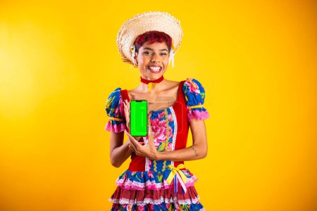Téléchargez les photos : Femme brésilienne avec des vêtements de festa junina. Arraial, fête de Saint-Jean. montrant smartphone, avec écran vert. - en image libre de droit