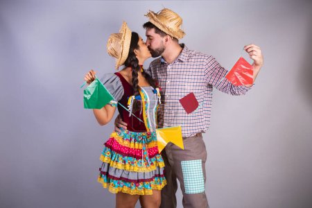Téléchargez les photos : Couple brésilien, vêtements de festa junina, arraial, festa de so joo. petit ami et petite amie. tenant des drapeaux, embrassant - en image libre de droit