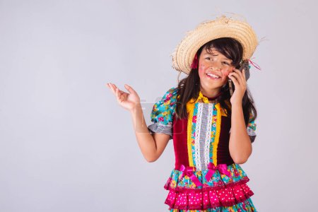Téléchargez les photos : Enfant, fille brésilienne, avec des vêtements de fête de juin, parlant sur un téléphone portable. - en image libre de droit