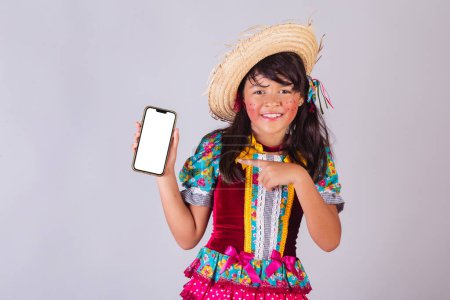 Foto de Niño, chica brasileña, con ropa de fiesta junina, mostrando la pantalla del teléfono inteligente. - Imagen libre de derechos
