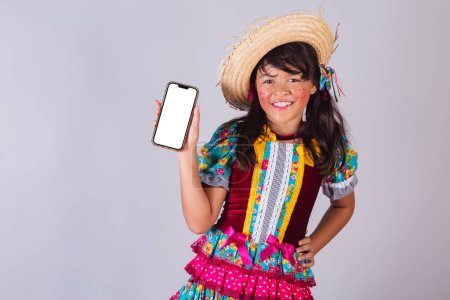 Téléchargez les photos : Enfant, fille brésilienne, avec des vêtements de fête junina, montrant l'écran du smartphone. - en image libre de droit