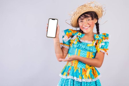 Foto de Niño, chica brasileña, con ropa de fiesta junina, mostrando la pantalla del teléfono inteligente. - Imagen libre de derechos