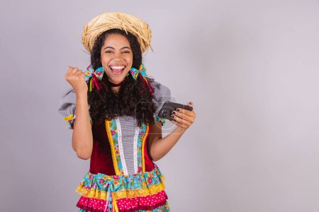 Foto de Negra, brasileña, vestida con ropa de fiesta de junio, fraternización en nombre de So Joo Arraial. con smartphone - Imagen libre de derechos
