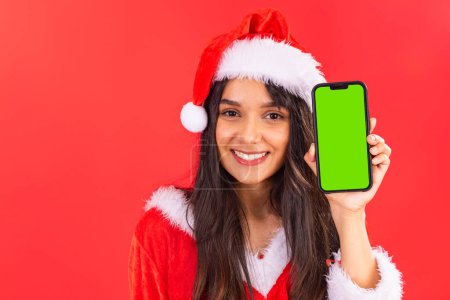 Photo for Brazilian teenage woman, wearing Christmas clothes, showing smartphone with green screen in chroma key, mobile advertisement, app. - Royalty Free Image