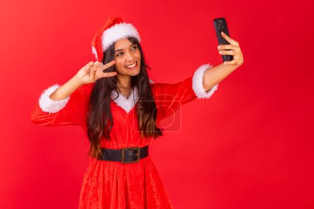 Photo for Brazilian teenage woman wearing Christmas clothes, smartphone taking selfie. - Royalty Free Image