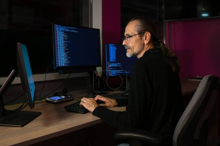 Téléchargez les photos : Un programmeur d'âge moyen avec des lunettes et une queue de cheval travaille avec du code sur un lieu de travail avec trois moniteurs dans un bureau sombre - en image libre de droit