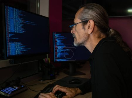 Téléchargez les photos : Un programmeur d'âge moyen avec des lunettes et une queue de cheval est en train de coder dans un bureau sombre - en image libre de droit