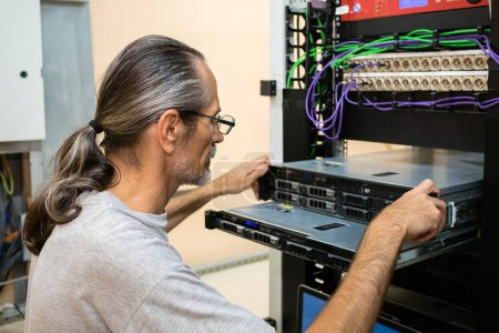 Photo for A middle-aged engineer with glasses and a ponytail maintains servers installed in a rack - Royalty Free Image