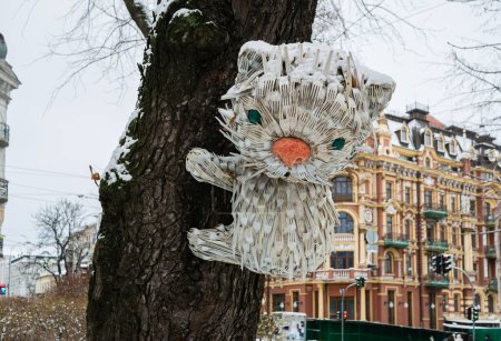 Foto de Kiev, Ucrania - 21 de enero de 2024: Gato cubierto de nieve hecho de tenedores de plástico desechables usados en un tronco de árbol en un viejo parque de la ciudad de invierno - Imagen libre de derechos