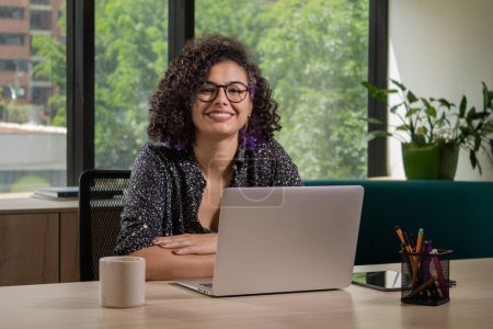 Portrait of a cheerful Latin woman with curly hair, wearing eyeglasses, sitting at a desk in a creative office. Using her laptop and looking at the camera with a confident smile.