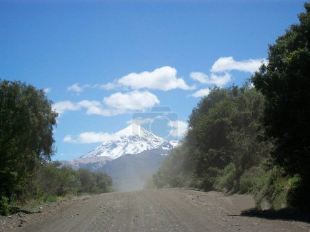 Dusty rural road surrounded by vegetation. In the background a snow peaked volcano with white clouds above.