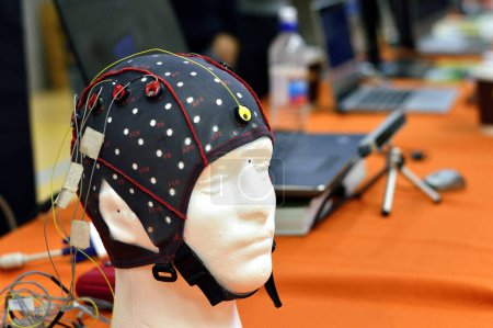 Téléchargez les photos : Bouchon de tête électroencéphalogramme (EEG) avec disques métalliques plats (électrodes) fixés à un modèle de tête en plastique blanc. Ordinateurs portables flous à l'arrière-plan. - en image libre de droit