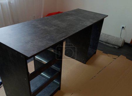 Auf dem Foto ist in der Mitte des Raumes ein schwarzer Tisch zu sehen, der während der Montage auf einer Papiermatte zur Hälfte gefaltet ist. Der Rahmen des neuen Tisches. Das Thema der selbstständigen Montage von Möbeln im Raum.
