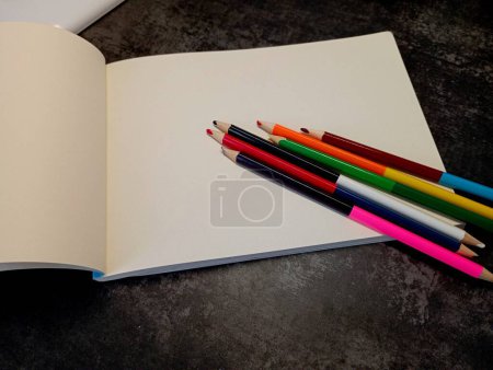 Double-sided colored pencils lie on an open drawing album with blank white pages. A drawing album and colored pencils are on the desktop