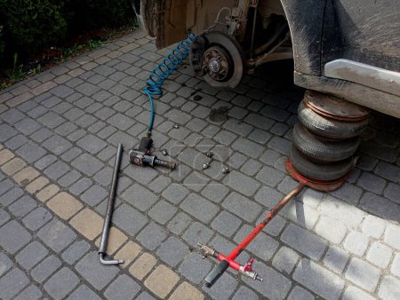 The front of the car is raised on a lift and with the wheel removed. There is a pneumatic wrench near the car and the bolts that hold the car wheel are lying on the cobblestones. The topic of seasonal tire replacement