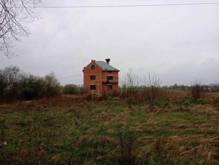 A large brick two-story house stands alone in a field on the chimney of which storks have built a large nest. The topic of architecture and construction and abandoned new unfinished buildings.