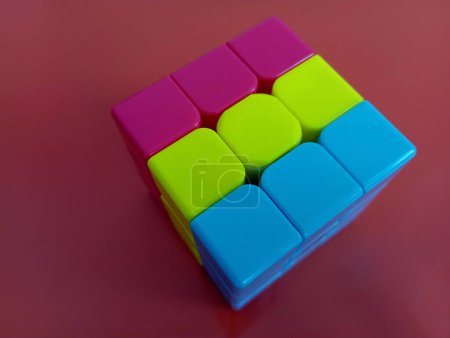 Téléchargez les photos : Un cube Rubik trois couleurs de couleurs jaune, bleu et rouge est empilé sur un fond bordeaux foncé. un jouet pour le développement de la pensée logique et de la motricité manuelle. - en image libre de droit