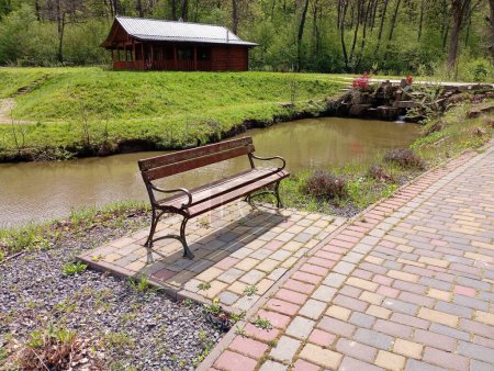 Eine Bank im Park auf einer Kopfsteinpflasterplattform in der Nähe eines Fußweges im Hintergrund eines Hauses und eines mit Wasser gefüllten Reservoirs. Ein schöner Ort zum Entspannen am Wasser im Park.