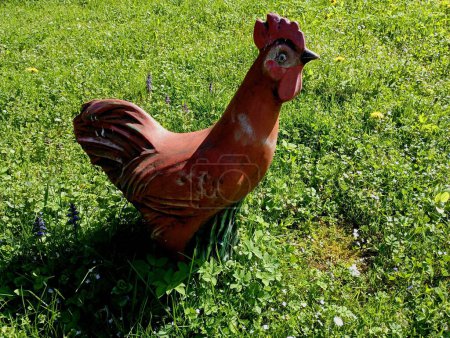Skulptur eines tönernen Hahns auf einem grünen Rasen. Ein brauner Lehmhahn streckte seinen Kopf in die Höhe und legte sich auf das grüne Gras auf dem Rasen.