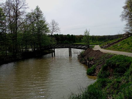 Foto de Una hermosa pasarela de madera a través de un estrecho canal que fluye entre la orilla del lago y una pequeña isla en la que crecen los árboles. Parque y embalses con agua. - Imagen libre de derechos
