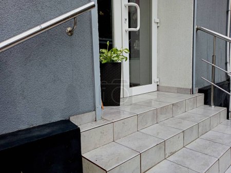 Escaliers avec balustrades métalliques fixés dans le mur sont doublés de carreaux de céramique menant à la porte en plastique de verre blanc avant de la maison, à côté de laquelle il y a un pot avec une plante.