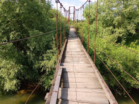 A wooden suspension bridge across a river on old rusty cables and a wooden crash surface made of old planks is laid across a river with clear water. A beautiful bridge over a reservoir for pedestrians.