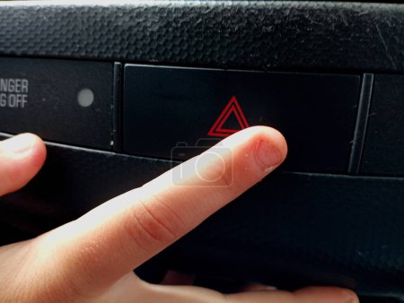 A person with his hand presses the button to turn on the emergency side lights indicated by a red triangle on the black instrument panel in the car.