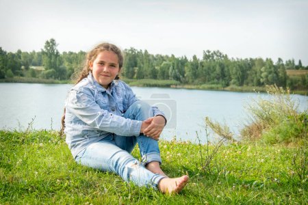 Téléchargez les photos : En été, une fille vêtue d'une veste en jean et d'un jean est assise sur la plage au bord de la rivière.. - en image libre de droit