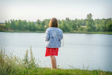 Téléchargez les photos : En été, une fille vêtue d'une veste en jean et d'une robe rouge se tient sur la rive de la rivière. - en image libre de droit